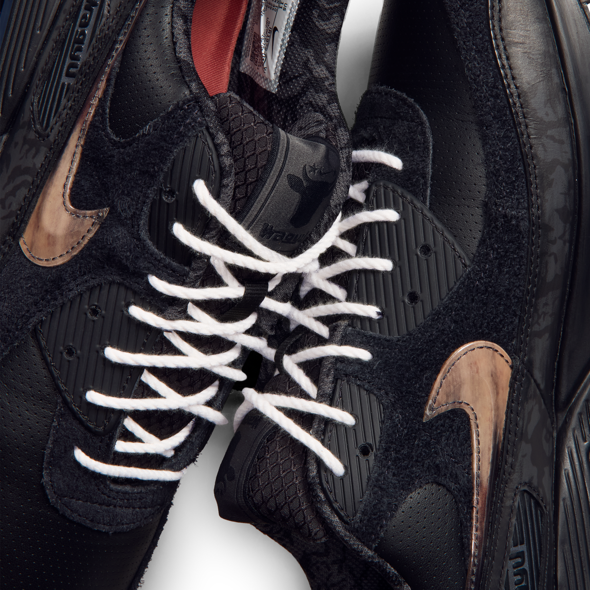Close-up of a black Nike sneaker with white laces, black textured overlays, and metallic bronze swooshes on a white background.