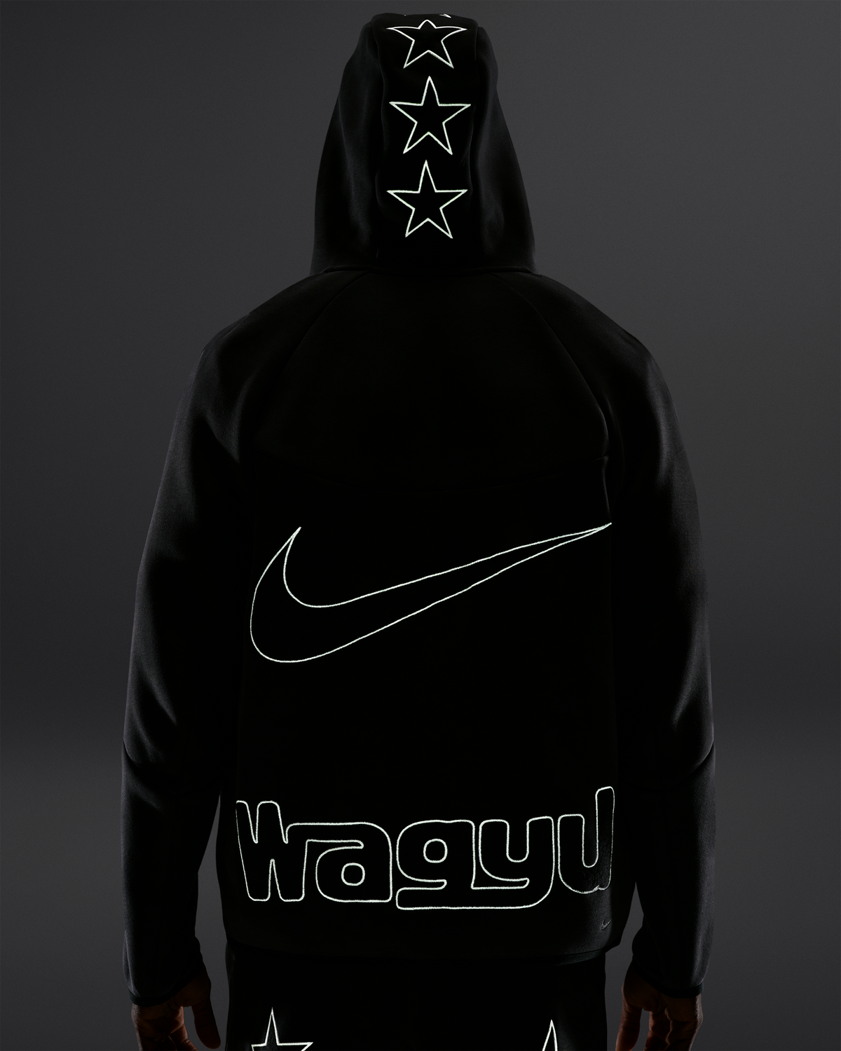 A person shown from the back wearing a black hoodie with a large white outlined Nike swoosh across the back, three outlined stars down the hood, and stylized text reading 'Wagyu' near the lower back. The background is a dark gray studio backdrop.